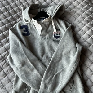 Grå hoodie från Polo Ralph Lauren - Säljer en stilren grå hoodie från Polo Ralph Lauren med broderad logga och nummerdetalj. Tröjan har en klassisk känguruficka och justerbar huva med snören. Perfekt för en avslappnad stil.