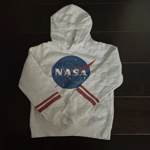 Vit hoodie med NASA-tryck från H&M - Säljer en cool vit hoodie från H&M med det ikoniska NASA-trycket på framsidan. Tröjan har en klassisk huva och långa ärmar med röda ränder vid ärmsluten. Perfekt för rymdentusiaster! 🚀