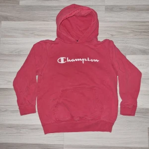 Röd hoodie från Champion - Säljer en röd hoodie från Champion med klassisk logga på bröstet. Tröjan har en stor ficka framtill (och några små fläckar) och en bekväm huva. Perfekt för en avslappnad stil. Passar för ungdomar i åldern 11-12 år.