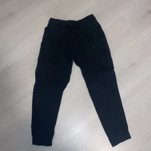 Svarta mjukisbyxor från Nike - Säljer ett par svarta mjukisbyxor från Nike. Byxorna har en bekväm passform med snörning i midjan och ribbade muddar vid bensluten. Perfekta för en avslappnad stil.