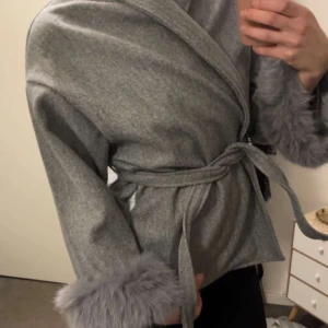 Grå kappa med pälsdetaljer - Snygg grå kappa med knytbälte och pälsdetaljer vid ärmsluten. Perfekt för kyliga dagar när du vill vara både varm och stilren.