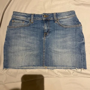 Blå jeanskjol från Tommy Hilfiger - Snygg blå jeanskjol från Tommy Hilfiger med fransig nederkant och klassisk femficksdesign. Perfekt för en avslappnad stil. Den har en knapp och dragkedja framtill.