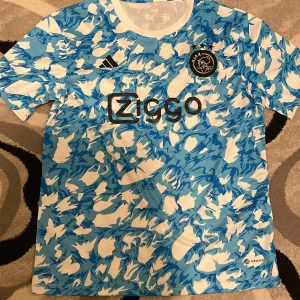 Blå och vit Ajax fotbollströja från Adidas - Säljer en cool blå och vit mönstrad Ajax fotbollströja från Adidas. Tröjan har korta ärmar och ett unikt mönster med klubbens emblem på bröstet. Varan är helt ny och ser väldigt fräsch ut.