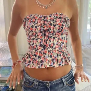 Blommig axelbandslös topp - Säljer en söt blommig axelbandslös topp med volangdetaljer. Toppen har en färgglad design, perfekt för sommaren🩷🩷