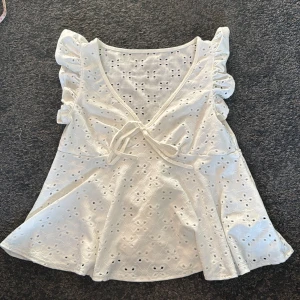 Vit blus med spetsdetaljer - Charmig vit blus med spetsdetaljer och volangärmar. Blusen har en v-ringad design med knyt framtill och ett fint broderat mönster. Perfekt för en somrig look! Använd fåtal gånger!