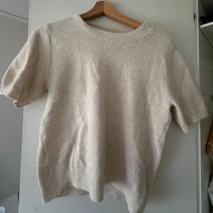 Beige stickad t-shirt från H&M - Mysig beige stickad tröja med korta ärmar och rund halsringning. Perfekt för en avslappnad stil. Tröjan har ribbade kanter vid ärmslut och nederkant.