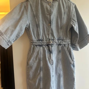 Jeansklänning från NORR med dragkedja - Säljer en snygg jeansklänning från NORR i ljusblå denim. Klänningen har knappar framtill, dragkedja i sidan och fickor. Modellen har halvlånga ärmar och markerad midja för en cool siluett. Perfekt till sneakers eller boots.