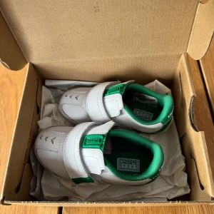 Vita sneakers från Lacoste - Snygga vita sneakers från Lacoste med gröna detaljer och kardborreband för enkel på- och avtagning. Skorna har en sportig design med märkets logotyp på både tungan och hälen. Supergulliga!