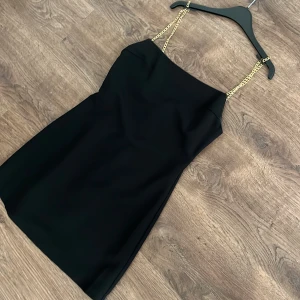 Svart klänning med kedjeband från Bik Bok - Snygg svart klänning från Bik Bok med guldfärgade kedjeband som axelband. Klänningen har en elegant och stilren design, perfekt för en kväll ute. Den är kort och har en smickrande passform.
