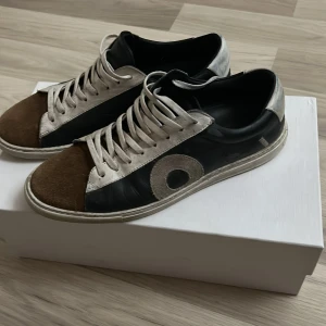 Svart och brun sneaker från Oliver Cabell - Snygga sneakers från Oliver Cabell i svart och brunt med vita detaljer. Skorna har en unik design med en stor cirkel på sidan och snörning framtill. Perfekta för en stilren look.