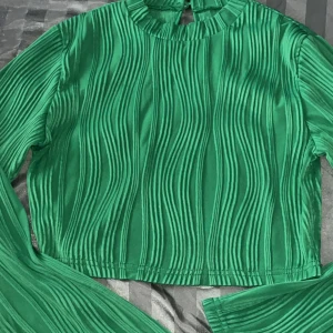 Grön ribbad topp - Snygg grön ribbad topp med långa ärmar och ett unikt vågigt mönster. Perfekt för att sticka ut med stil. Toppen har en hög hals och en modern look.