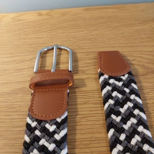 Flätat bälte med läderdetaljer - Snyggt flätat bälte i svart, grått och vitt med bruna läderdetaljer. Bältet har ett silverfärgat spänne och en unik design som ger en stilren look. Perfekt för att ge din outfit det lilla extra.