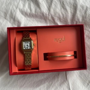 Guldfärgad klocka med länkarmband från Regal - Elegant guldfärgad klocka från Regal med fyrkantig urtavla och romerska siffror. Klockan har ett länkarmband och kommer i en presentförpackning tillsammans med ett matchande armband. Perfekt för att ge en lyxig touch till din stil.