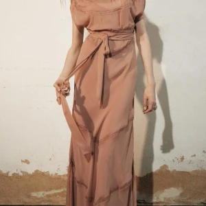 Beige långklänning med knytband - Elegant beige långklänning med knytband i midjan. Klänningen har en mjuk och flytande design med subtila detaljer som ger en romantisk känsla. Perfekt för speciella tillfällen.