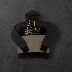 Grå och svart hoodie från Superdry - Säljer en cool hoodie från Superdry i grått och svart med limegröna detaljer. Den har en stor logga på bröstet och en justerbar huva med vita snören. Perfekt för en avslappnad stil.