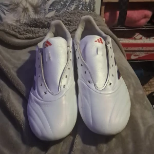Vita Adidas fotbollsskor - Säljer ett par vita Adidas fotbollsskor med blå och röda detaljer. Skorna har en stilren design med de klassiska tre ränderna på sidan. Perfekta för fotbollsplanen. Änvändt I 3 matcher💫perfekt skick 💥💫 ny pris 1200