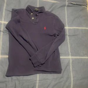 Mörkblå pikétröja från Polo Ralph Lauren - Säljer en stilren mörkblå pikétröja från Polo Ralph Lauren med klassisk krage och knappar. Tröjan har ett broderat rött logotypmärke på bröstet och långa ärmar. Perfekt för en avslappnad men ändå elegant look.