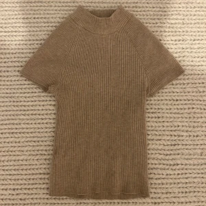Beige ribbad kortärmad tröja - Säljer en stilren beige ribbad tröja med korta ärmar. Perfekt för en avslappnad look. Tröjan har en högre krage och är i ett mjukt material. Aldrig använd