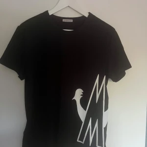 Svart t-shirt från Moncler - Snygg svart t-shirt från Moncler med ett stort vitt tryck på framsidan och Moncler-logga på ärmen. Perfekt för en stilren look. Storlek:M men passar S/Xs          Nypris: 4000