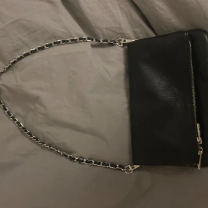 Svart axelväska med kedja - Snygg svart axelväska i skinn med en stilren kedja som axelrem. Väskan har en praktisk dragkedja framtill för enkel åtkomst till dina saker. Perfekt för att ge en elegant touch till din outfit.