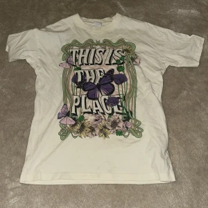 Vit t-shirt med fjärilstryck - Säljer en vit t-shirt med ett stort tryck på framsidan som har texten 'This is the place' omgiven av lila fjärilar och blommor. T-shirten har korta ärmar och en avslappnad passform. Perfekt för en avslappnad stil.