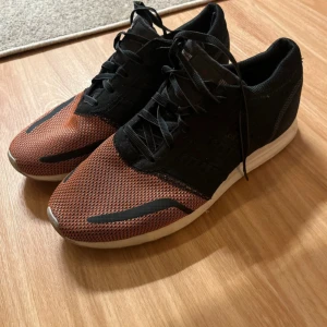 Adidas skor  - Snygga sneakers i svart och orange med snörning. Skorna har en vit sula och en sportig design, perfekt för en aktiv livsstil.