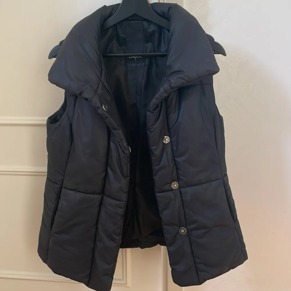 Säljer en svart dunväst från Lindex med knappar och en hög krage. Perfekt för kyligare dagar när du vill hålla värmen utan att bära en jacka. Västen har en stilren design och passar till många olika outfits.. Takit.