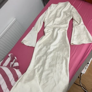 Vit långklänning med mönster - Elegant vit långklänning med subtilt mönster och vida ärmar. Klänningen har en snörning i ryggen som ger en fin passform. Perfekt för speciella tillfällen.