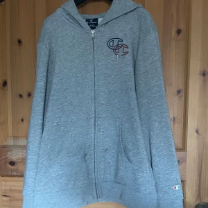 Grå hoodie från Champion - Säljer en grå hoodie från Champion med dragkedja och broderad logga på bröstet. Tröjan har långa ärmar och en bekväm passform. Perfekt för en avslappnad stil.