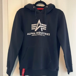 Svart hoodie från Alpha Industries - Säljer en svart hoodie från Alpha Industries med deras ikoniska logga tryckt på framsidan. Hoodien har en klassisk design med dragsko i huvan och en praktisk magficka. Perfekt för en avslappnad stil.