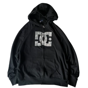 Svart hoodie från DC - Säljer en svart hoodie från DC med dragkedja och stort tryck på framsidan. Perfekt för en avslappnad stil. Den har en praktisk huva och fickor framtill.