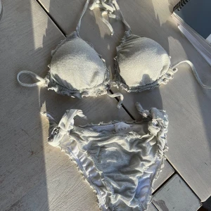 Grå silvrig glittrig bikini med volangdetaljer - Glittrig Snygg grå silvrig bikini med volangdetaljer och knytband. Toppen har vadderade kupor och knytning i nacken och ryggen. Trosorna har också knytband på sidorna för justerbar passform.