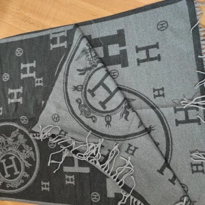 Grå och svart halsduk med H-mönster - Snygg och stilren halsduk i grått och svart med ett elegant H-mönster. Den har fransar i ändarna och ett stort cirkulärt emblem med hästmotiv. Perfekt för att ge en touch av lyx till din outfit.