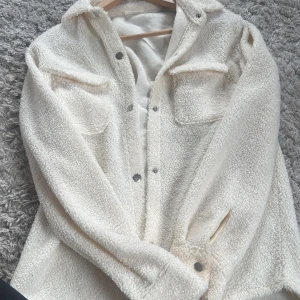 Vit teddyjacka från KappAhl - Mysig vit teddyjacka från KappAhl med knappar framtill och två fickor i fram. Perfekt för kyliga dagar med sin fluffiga och varma design. Jackan har långa ärmar och en klassisk krage. Utan defekter