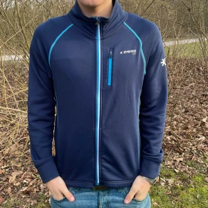 Mörkblå fleecejacka från Everest - Säljer en mörkblå fleecejacka från Everest med dragkedja och turkosa detaljer. Jackan har långa ärmar och en hög krage, perfekt för kyliga dagar. Den är både stilren och funktionell, med en broderad logga på bröstet och ärmen.