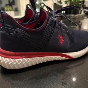 Polo skor - Snygga mörkblå sneakers från U.S. Polo Assn. med röd logga och detaljer. Skorna har en vit sula och snörning framtill. Perfekta för en sportig look.