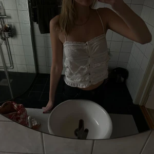Vit linnetopp - Säljer en söt vit linnetopp med spetsdetaljer och knappar framtill. Toppen har smala axelband och en lätt croppad passform, perfekt för varma dagar.