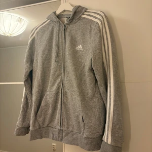 Grå hoodie från Adidas - Säljer en grå hoodie från Adidas med dragkedja och klassiska vita ränder längs ärmarna. Hör av er vid funderingar eller om ni vill ha bilder med hur plagget sitter på mig. Titta gärna in mina andra plagg!