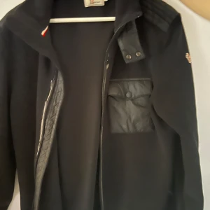 Svart moncler cardigan  - Snygg svart ull cardigan med dragkedja och en praktisk bröstficka med knapp. Jackan har en diskret logga på ärmen 