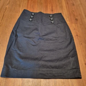 Svart kjol från Lindex - Snygg svart kjol från Lindex med dekorativa knappar framtill. Kjolen har en klassisk design och är perfekt för en stilren look. 