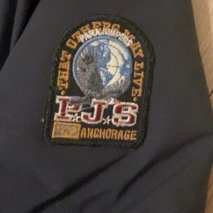 Svart pufferjacka från Parajumpers Lite tyg lossnat annars bra skick kan diskutera vid snabb affär  - Säljer en svart pufferjacka från Parajumpers med en cool patch på ärmen. Jackan har en dragkedja framtill och en huva för extra skydd. Perfekt för kyliga dagar.