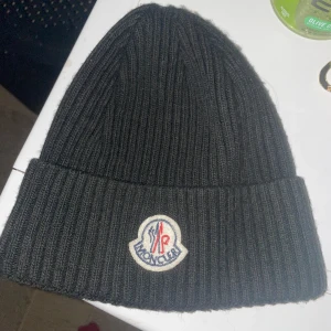Svart ribbad mössa från Moncler - Snygg svart ribbad mössa från Moncler med broderad logotyp framtill. Perfekt för att hålla värmen under kyliga dagar. Stilren design som passar till det mesta.den är för kvinna och män pris kan diskuteras