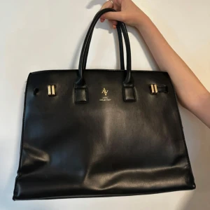 Svart handväska från AV Collection - Elegant svart handväska från AV Collection i skinn. Väskan har två handtag och stilrena detaljer i guld. Perfekt för en chic och sofistikerad look.