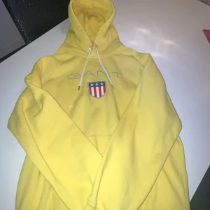 Gul hoodie Gant skön - Säljer en snygg gul hoodie från GANT med ett broderat emblem på bröstet. Hoodien har en justerbar dragsko i huvan och en stor ficka framtill. Perfekt för en avslappnad stil.