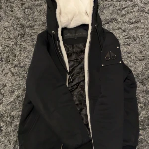 Svart jacka från Moose Knuckles (bunny) - Säljer en snygg svart jacka från Moose Knuckles med quiltad insida och mysig vit foder i huvan/(kan ta bort fodringen) Jackan har dragkedja och snörning vid huvan för extra stil och funktion. Perfekt för kyliga dagar! Där man vill se extra stilig utt 