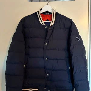 Mörkblå dunjacka från Gant - Säljer en snygg mörkblå jacka från Gant med en sportig look.  Den har en kontrasterande orange insida och ribbade detaljer vid krage och ärmslut. Perfekt när de e lite kallare. Nypris ca 2500. Storlek S