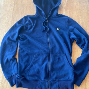 Lyle and Scott blå hoodie - Säljer en snygg blå hoodie från Lyle & Scott med dragkedja och huva. Den har praktiska fickor framtill och är perfekt för en avslappnad stil. Det finns ett minimalt hål på vänstra och högra armen men inget som märks