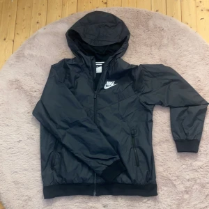 Svart vindjacka från Nike - Säljer en svart vindjacka från Nike med huva och dragkedja. Jackan har långa ärmar och två fickor framtill. Perfekt för blåsiga dagar och sportiga aktiviteter. Tillverkad i Vietnam. Den är bra använd men inget som syns, inga hål o.sv. storlek XL tydligen men sitter lika bra som storlek S
