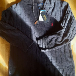 Svart stickad tröja från Polo Ralph Lauren - Säljer en snygg svart stickad tröja från Polo Ralph Lauren med det klassiska röda broderade logotypen på bröstet. Tröjan har en dragkedja vid halsen och ribbade muddar vid ärmslut och nederkant. Perfekt för en stilren look! (Ej originell)