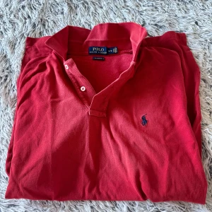 Röd pikétröja från Ralph Lauren - Säljer en klassisk röd pikétröja från Ralph Lauren med den ikoniska loggan broderad på bröstet. Tröjan har korta ärmar och en stilren krage med knappar. Perfekt för en avslappnad och stilren look.